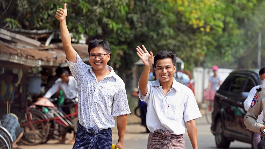 Reuters reporters Wa Lone and Kyaw Soe Oo wave to cameras as they walk down a dirt road carrying bright yellow and blue bags.