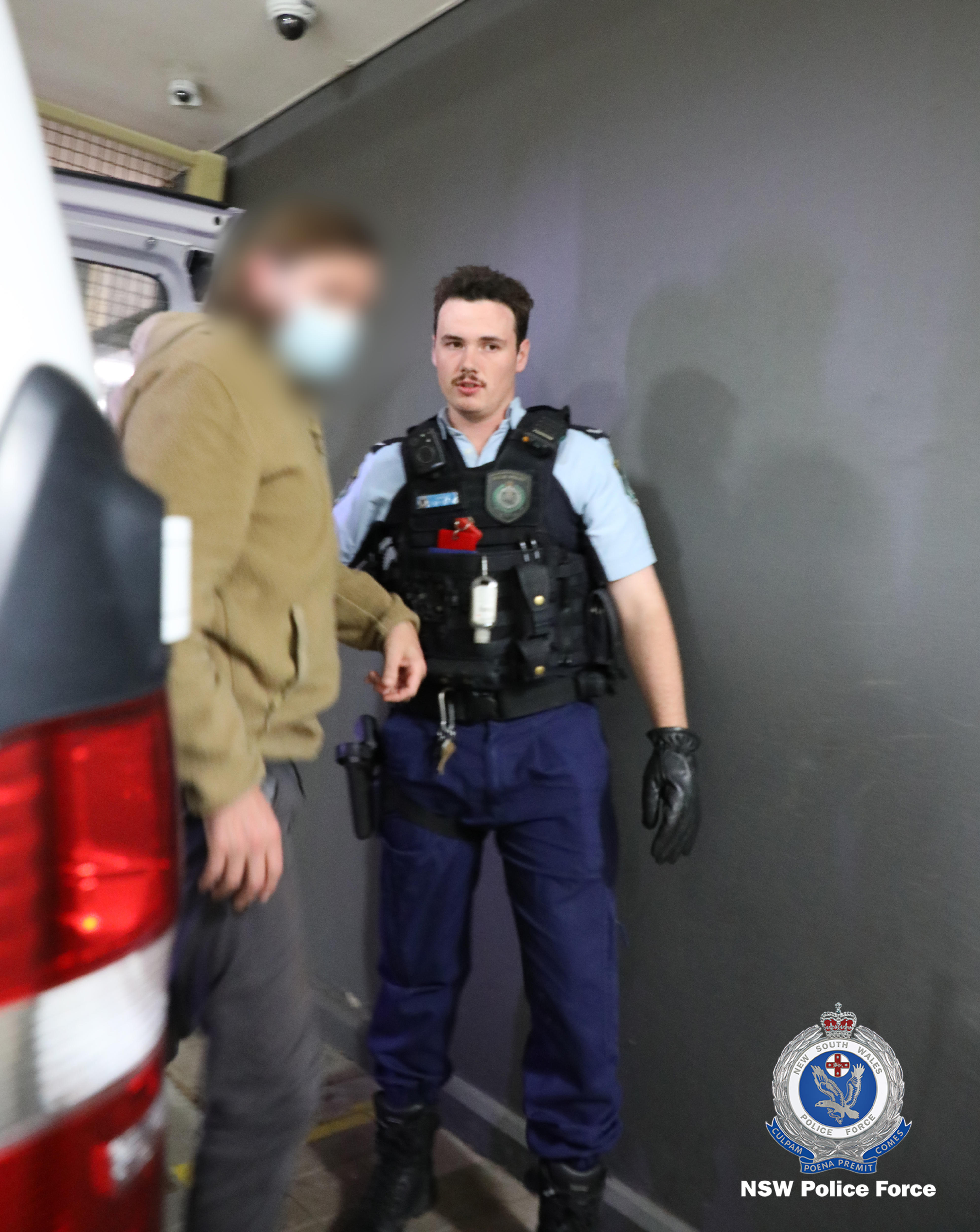 Man, face blurred being led out of the back of a car by a man wearing police uniform.