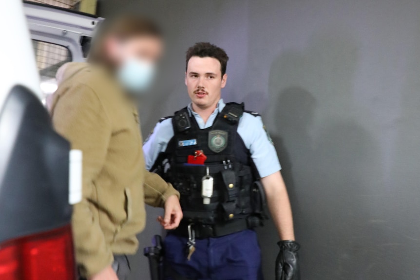 Man, face blurred being led out of the back of a car by a man wearing police uniform.