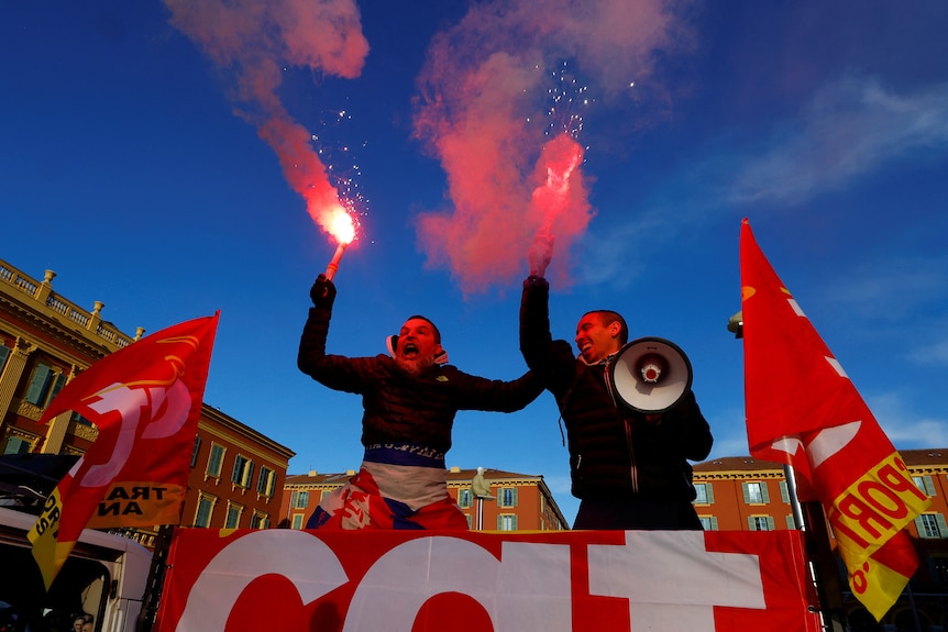 Two men fire pink flares and hold red flags in the air as they shout as part of a protest.