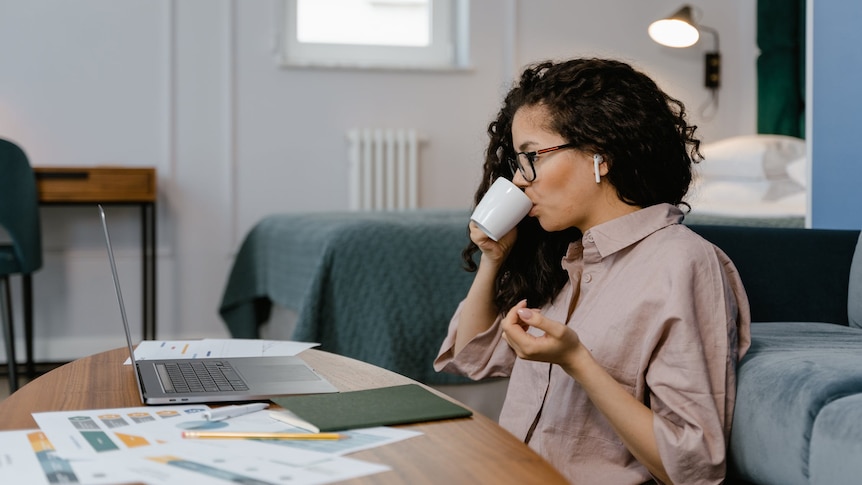 A woman sits at a coffee table, looking at her laptop, in story about tax time when working from home, have crypto investments.