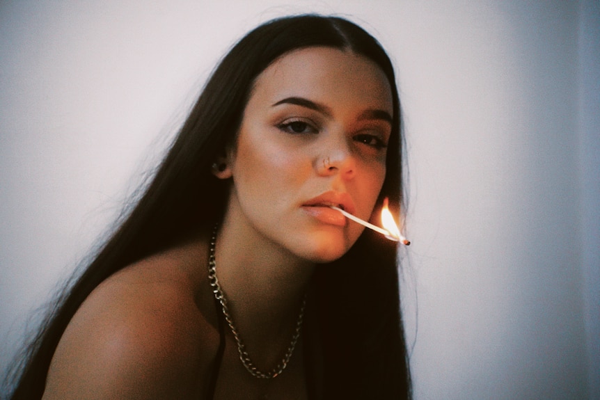 A young woman with long, dark hair, holding a burning match in her mouth.
