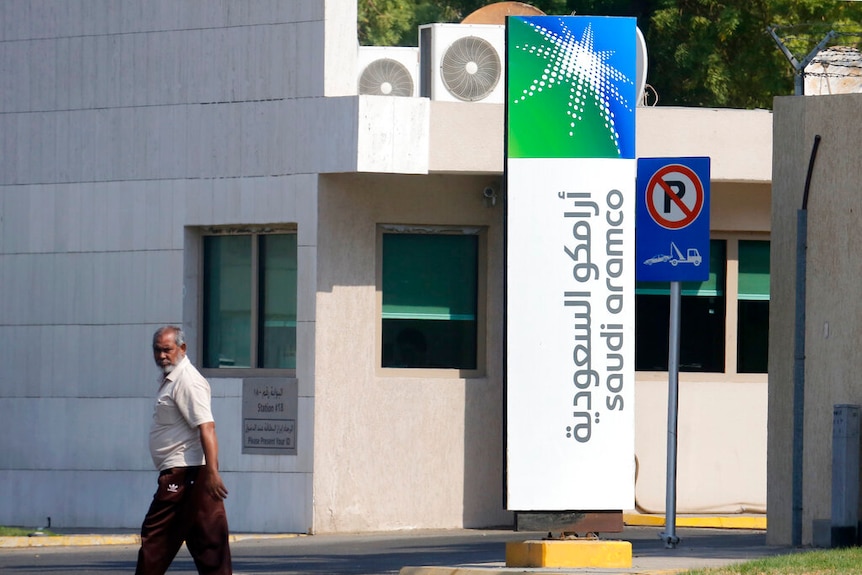 A man walks past the entrance to a compound with a large sign carrying the logo for Saudi Arabia's state oil company, Aramco.