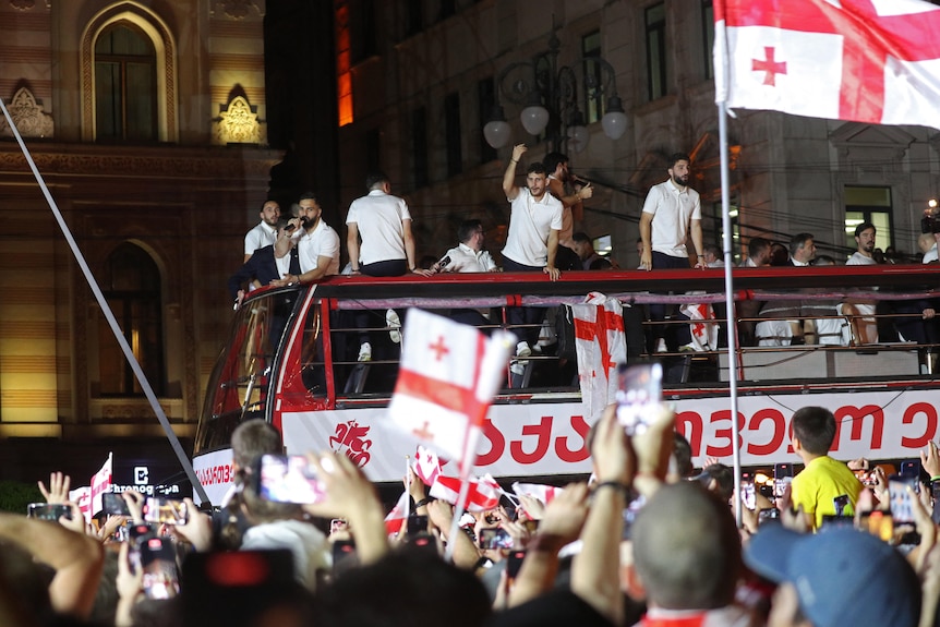 A group of footballers in white tops stand on an open-top bus waving at a big crowd of fans waving flags.
