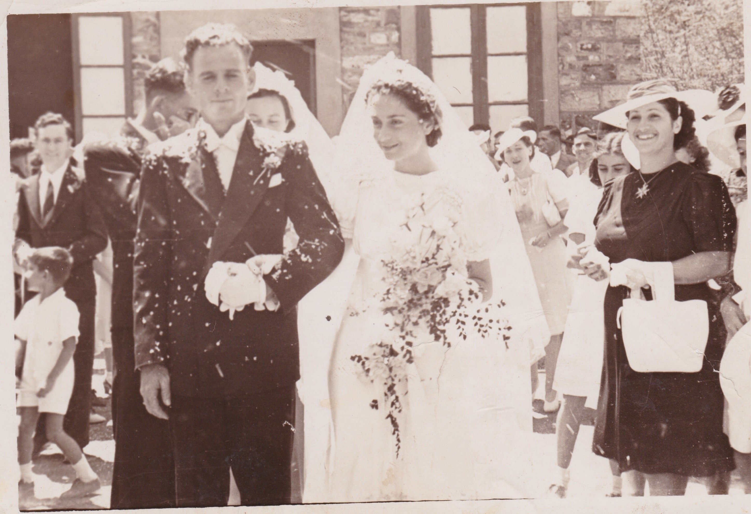 A 1940s sepia photo of a wedding, with a bride and groom outdoors, as others watch on