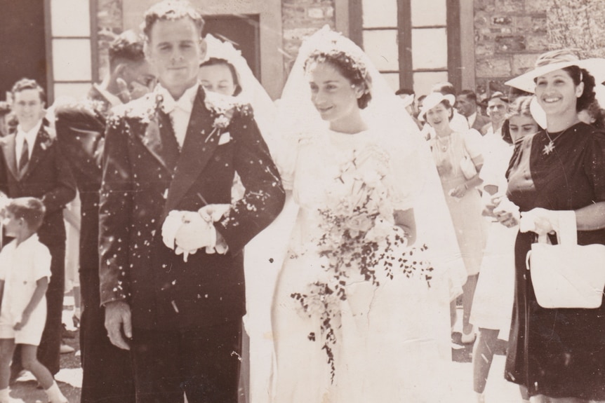 A 1940s sepia photo of a wedding, with a bride and groom outdoors, as others watch on