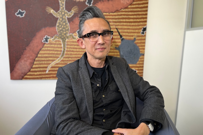 a man with glasses wearing a suit sits in front of a painting.