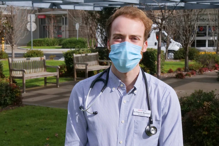 A man standing outside a hospital with a blue mask and a stethoscope around his neck