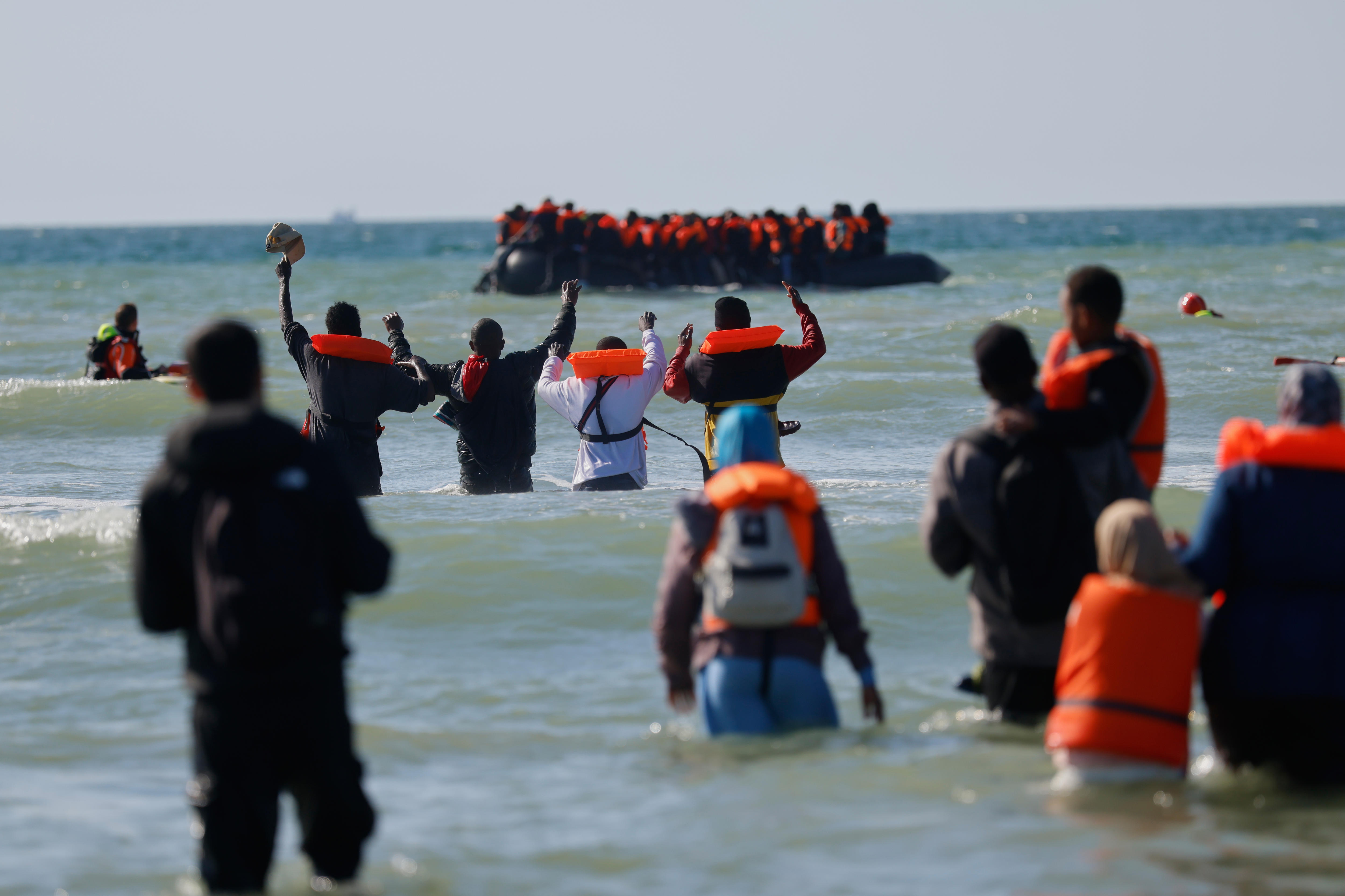 Groups of migrants wearing dark clothing and bright orange life vests wading in waist-deep water in front of a blurred boat.
