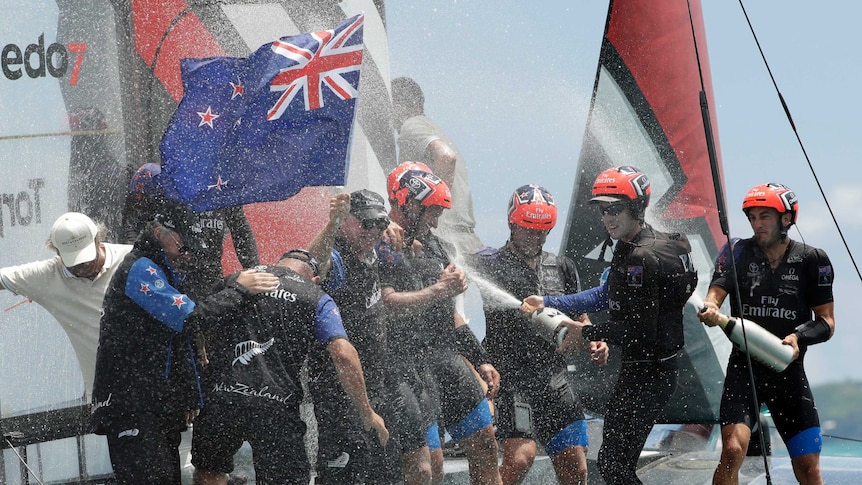 Two teammates spray oversized bottles of champagne over several other teammates while one holds the New Zealand flag