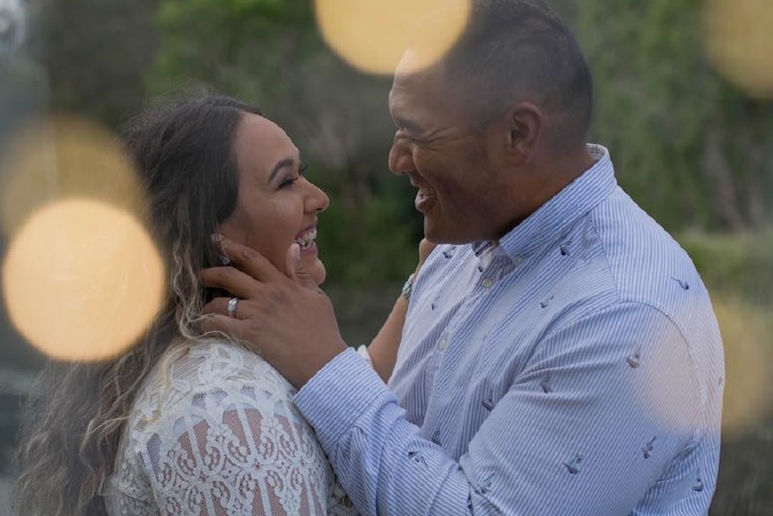 A woman with long dark hair and a man with short dark hair standing together holding each other and laughing