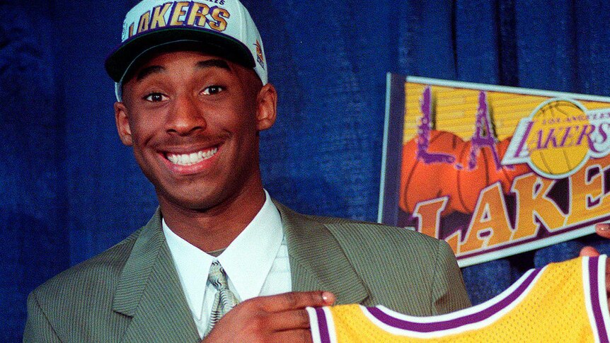 A smiling, young Kobe Bryant holds an LA Lakers jersey and wears a Lakers cap.