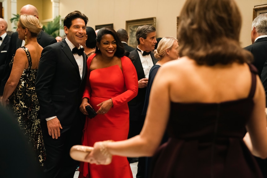 A middle-aged white man in a tuxedo stands with a middle-aged Black woman in a red floor-length dress at a formal event.