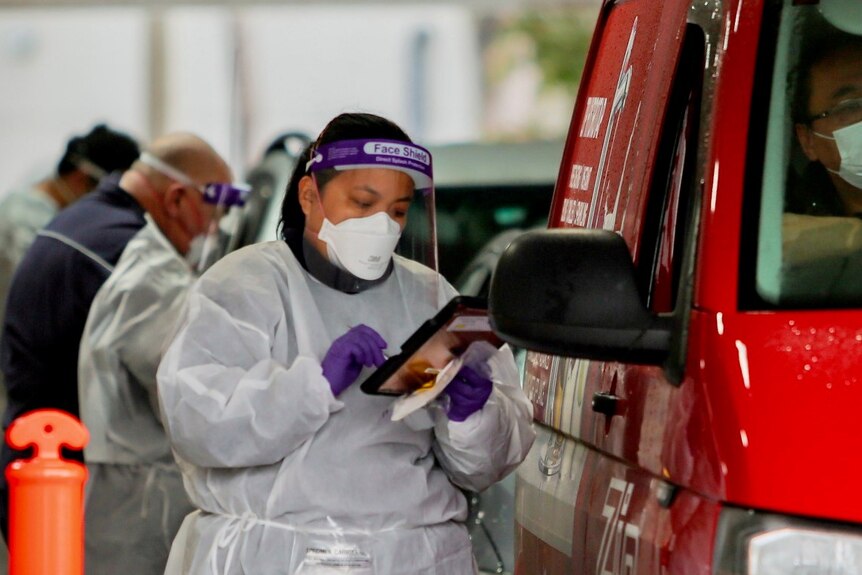 A healthcare worker in PPE near a driver in a red car at a testing site.