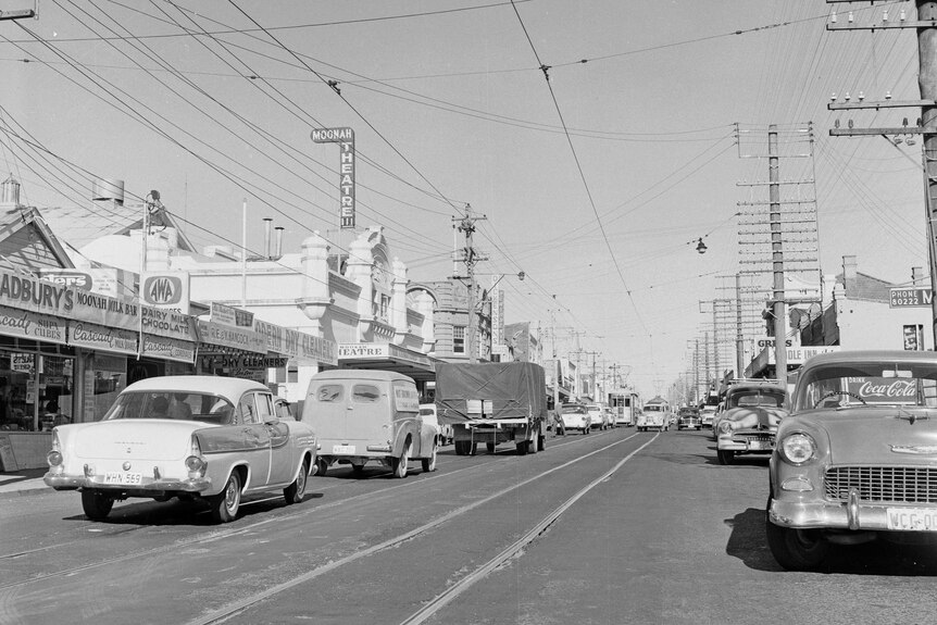 A black and white photo of a street with shops either side and old cars on the road