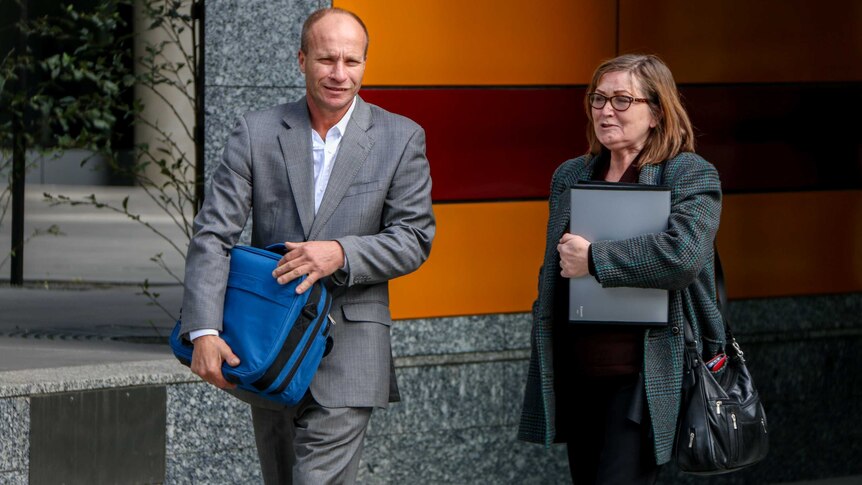 Christopher Somogy, wearing grey suit, white shirt and holding blue bag, walking next to woman wearing coat holding files.
