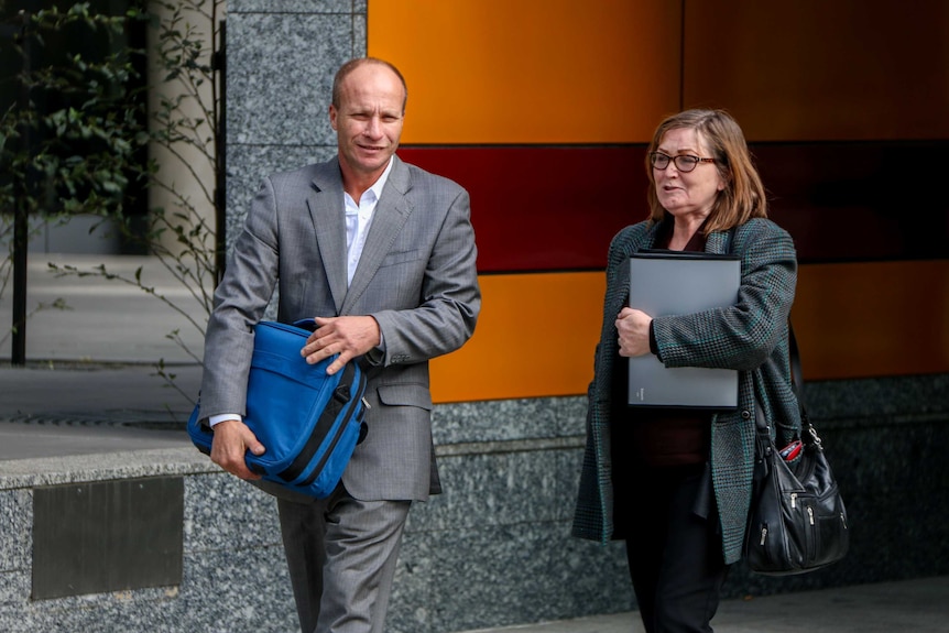 Christopher Somogy, wearing grey suit, white shirt and holding blue bag, walking next to woman wearing coat holding files.