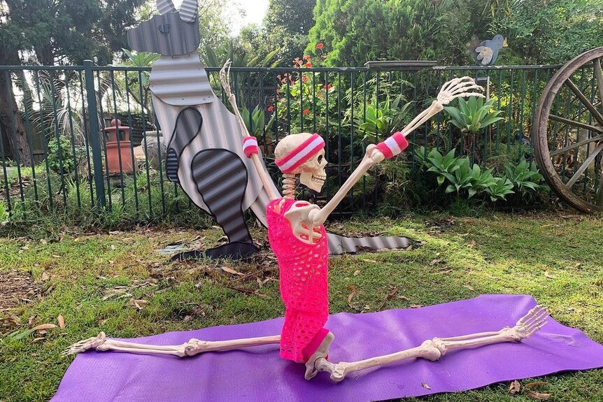 A skeleton doing the splits on a purple yoga mat on grass with its hands in the air.