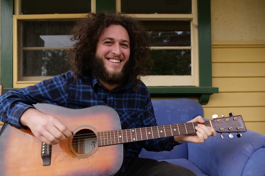 A man with a beard plays guitar on the couch.