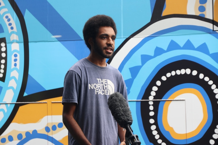 A young Indigenous man stands in front of a microphone. A large blue and yellow mural is behind him.