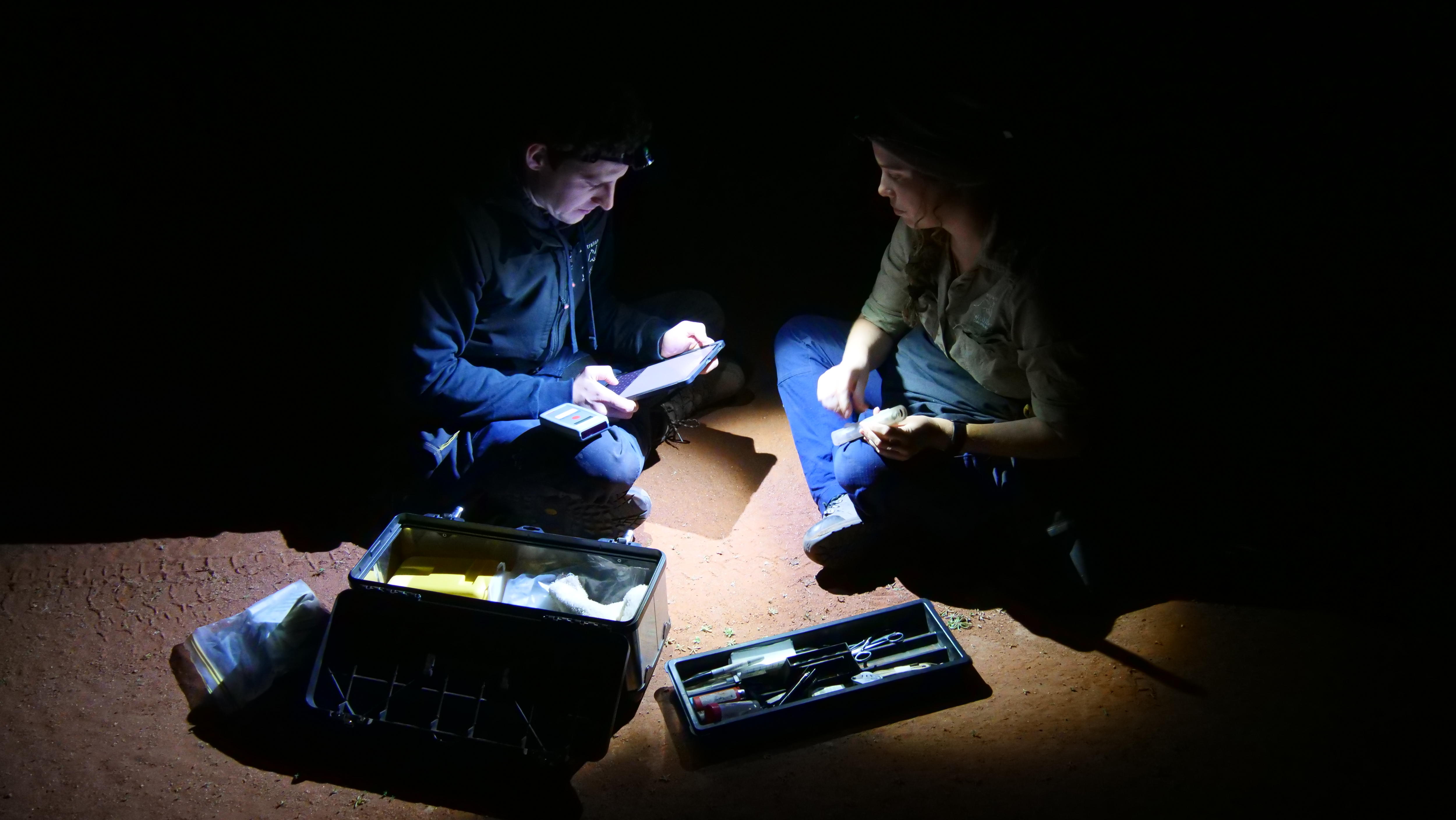 Two people sit on the ground, their head torches cast a circular glow over a hand held ipad, their laps and an open toolbox.