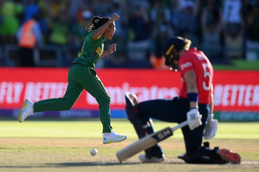 A South African bowler runs away with her arms raised in triumph as the ball sits next to an English batter on one knee.