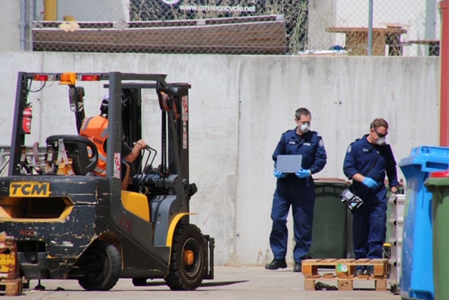Police officers in blue forensic overalls examine the site, while a fork lift operator works nearby.