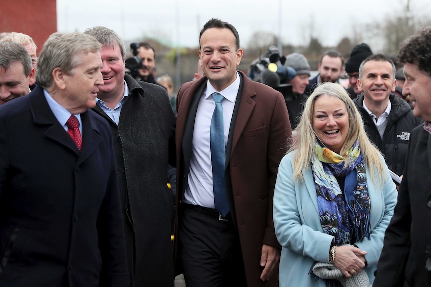 A man in a brown coat and blue tie smiles and walks among many men with woman in blue with long blonde hair beside him.