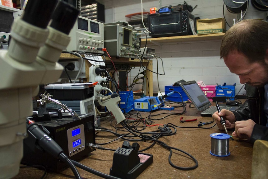 A man solders, surrounded by electronics equipment
