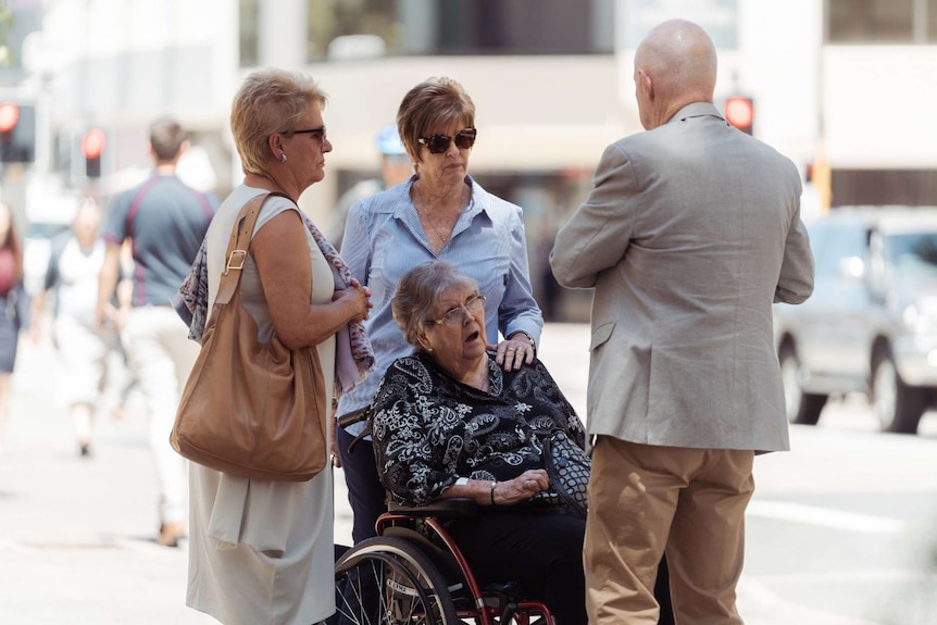 Jenny Rimmer in a wheelchair on a city street, surrounded by two other woman and a man in a light brown suit.