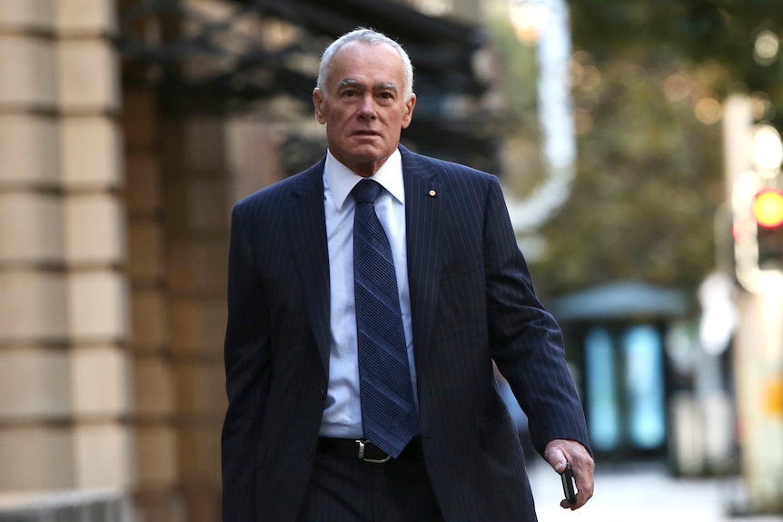 A mid-shot of John Poynton walking outdoors in the Perth CBD wearing a suit and tie.