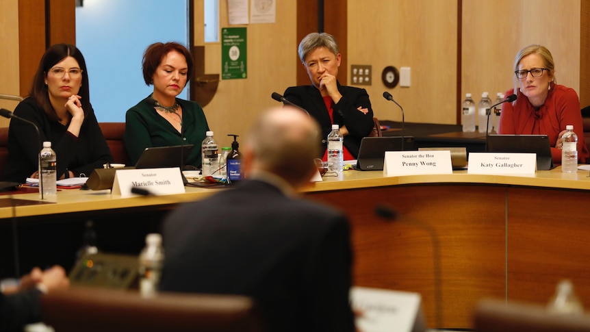 Kitching and Wong look serious as they stare at a man, whose back is facing the camera.