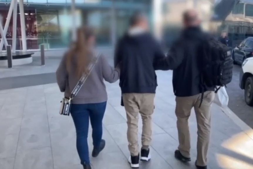 Seen from behind, a man is escorted by two plain-clothed police officers at Canberra Airport.