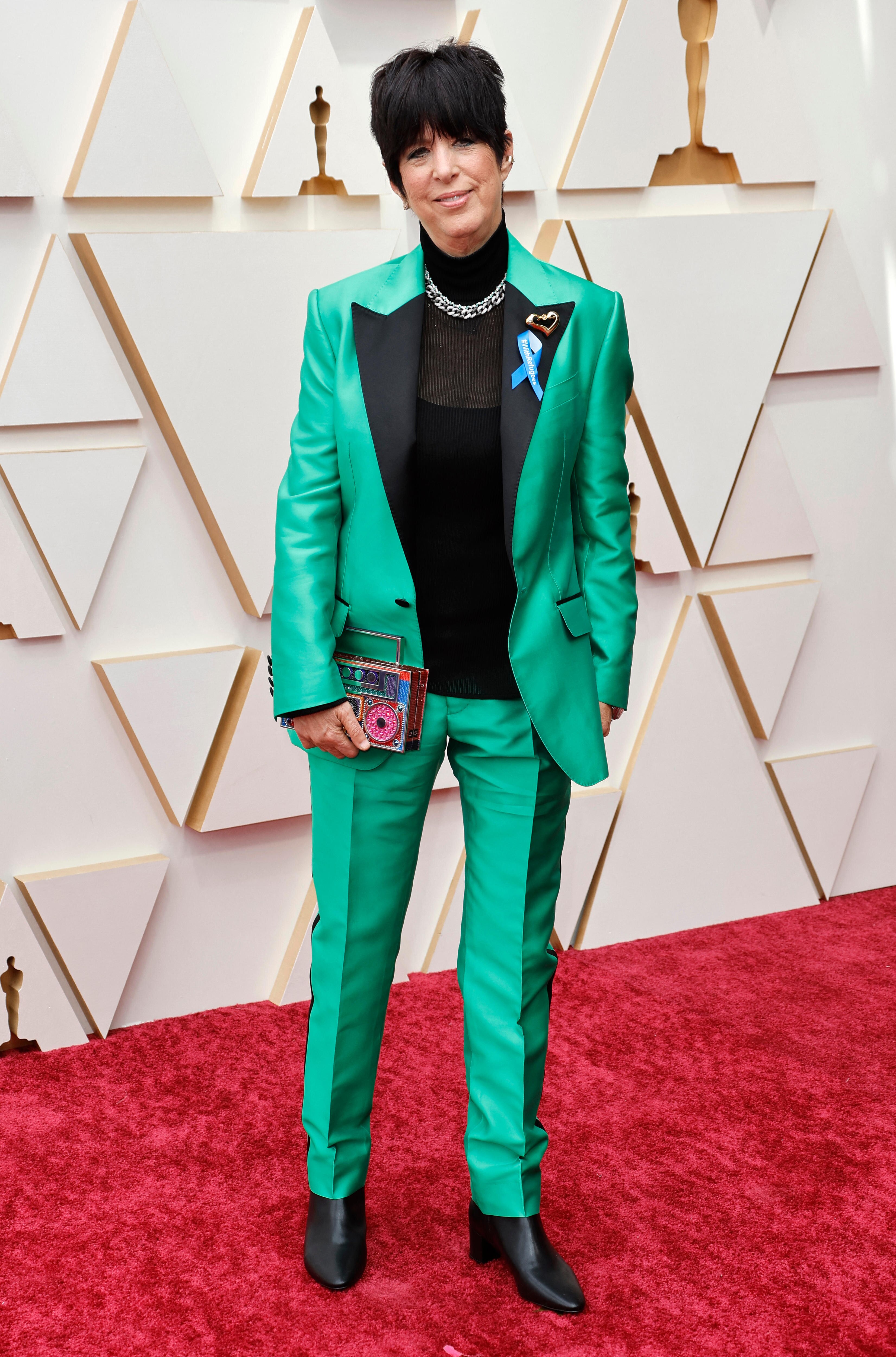 Musician Diane Warren stands on a red carpet, wearing a turquoise suit with a blue ribbon on its lapel.