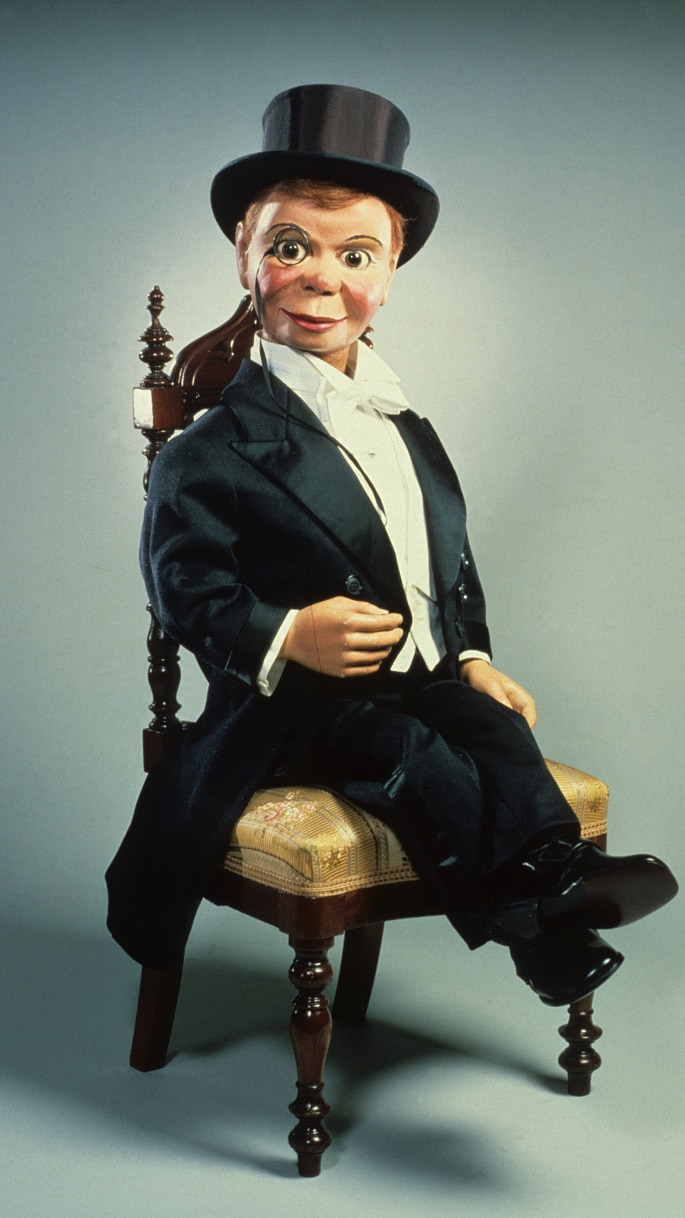 A ventriloquist dummy sits on a chair wearing a monocle