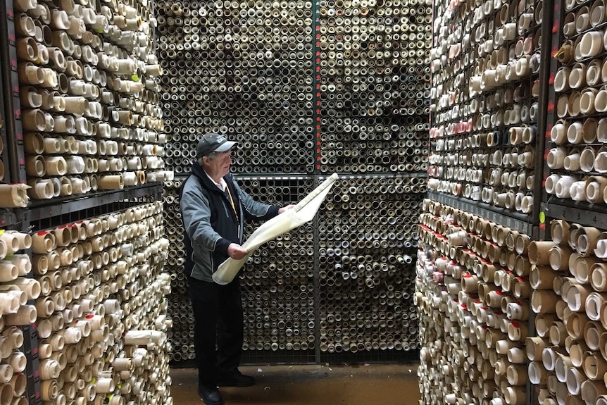 A man standing by shelves full of manufacturing plans, opens a roll of plans and looks at it