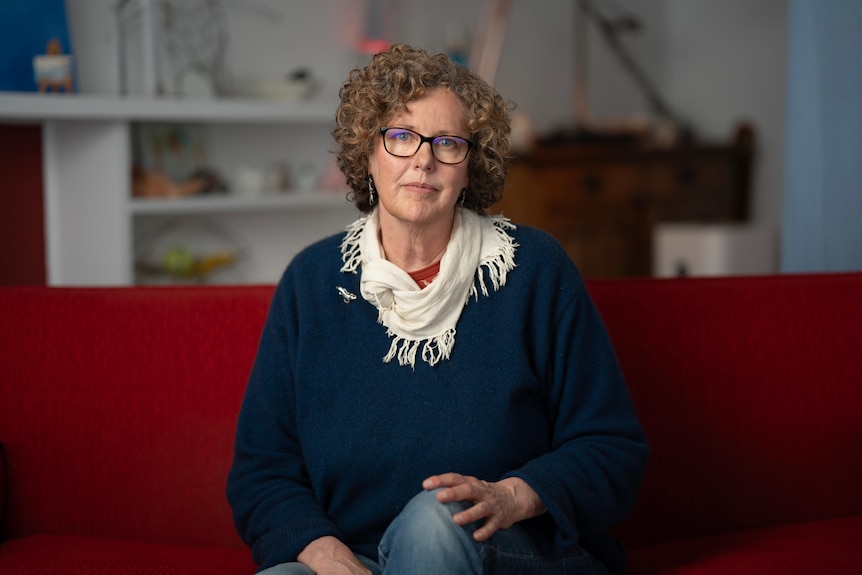 A woman wearing a blue jumper, white scarf sits on a red couch. 