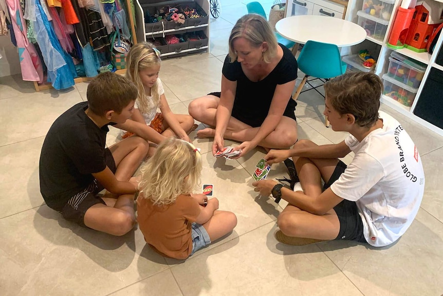 A mother and her four children playing cards while sitting on the floor of their kitchen