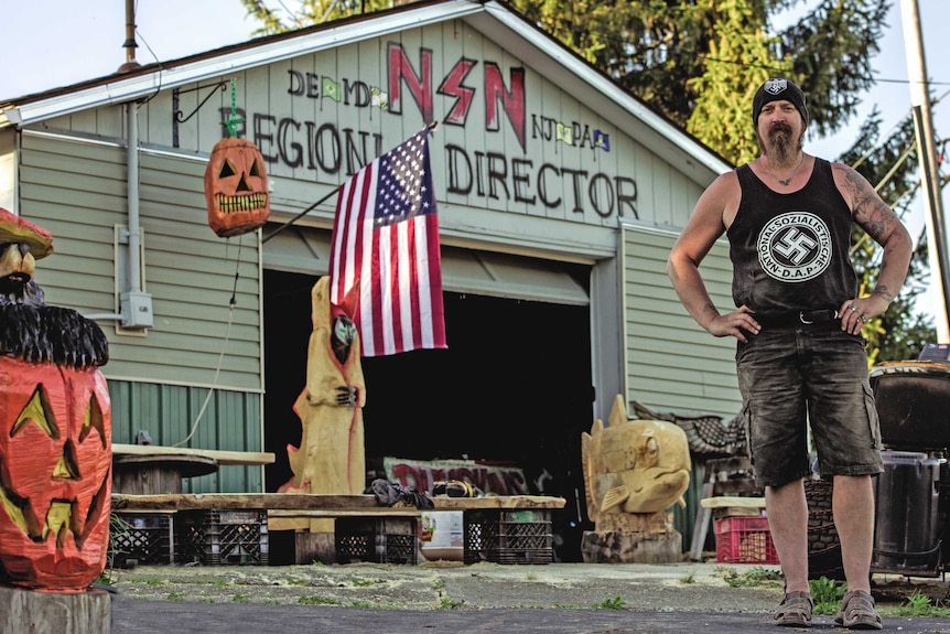A tattooed man in a singlet bearing a swastika stands in front of a workshop