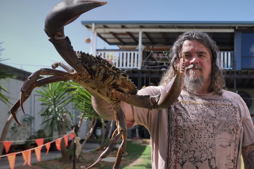 Smiling man with shoulder-length hair, grey beard holding a crab with its pincers out. House, small palm tree behind, blue sky.
