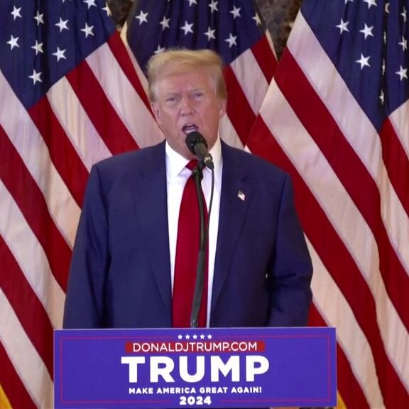 Blonde haired, tanned Donald Trump stands at podium in front of multiple American flags. 
