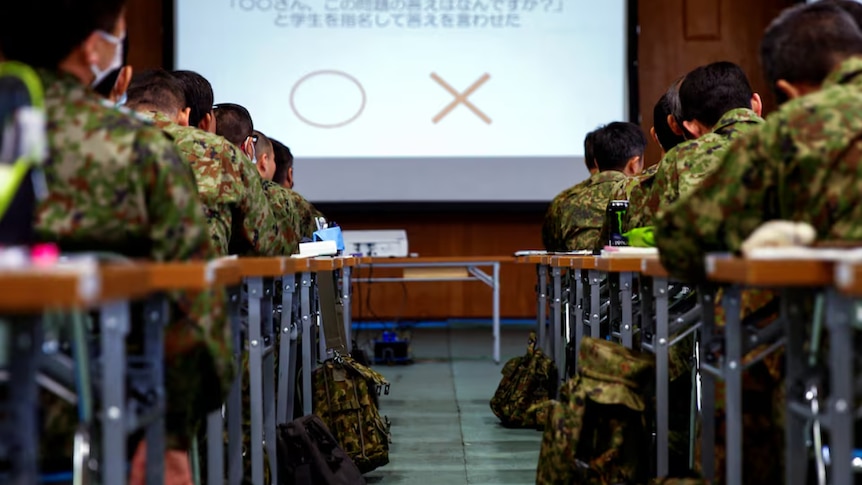 Japanese defence soldiers sit at tables in rows while looking at a presentation on a screen