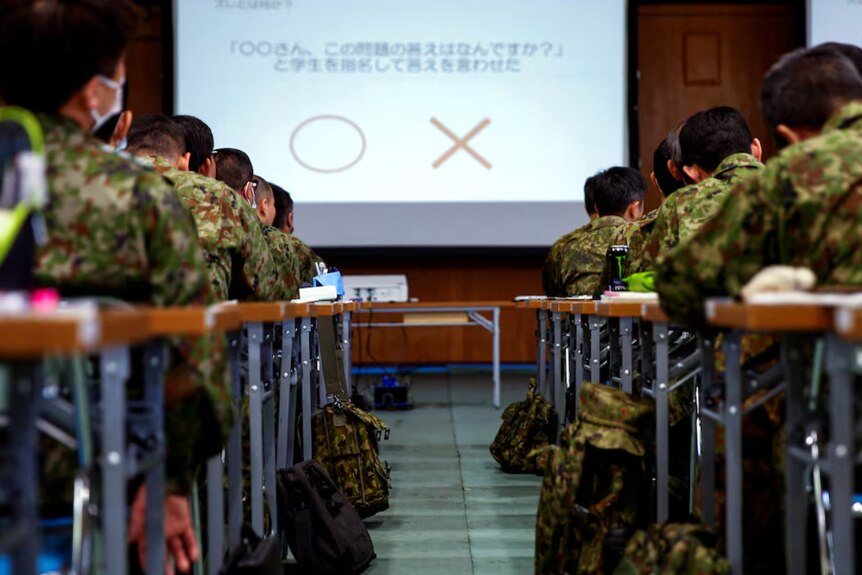 Japanese defence soldiers sit at tables in rows while looking at a presentation on a screen