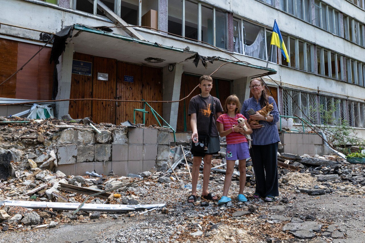 A woman and two young children standing outside a damaged building, looking at the camera