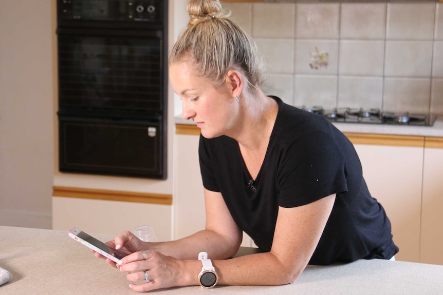 A woman leans on a kitchen bench scrolling on her mobile phone.