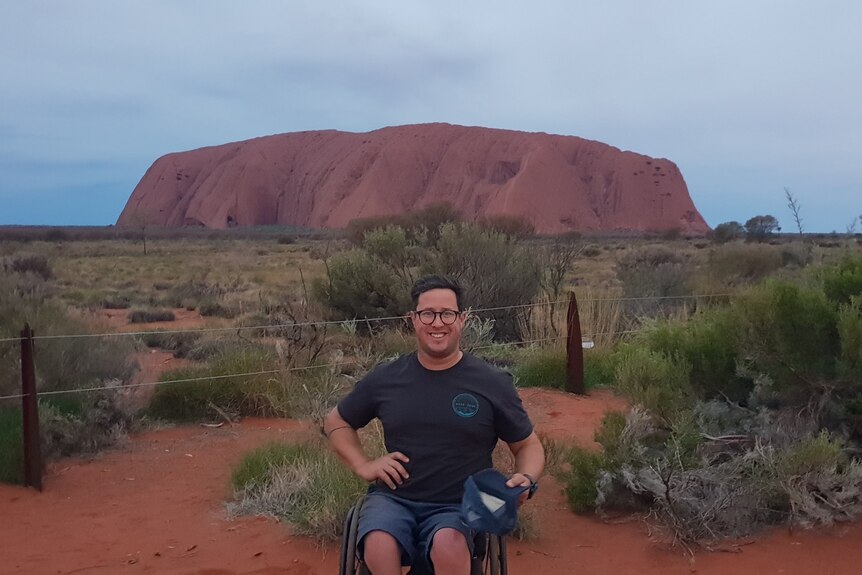 A man in a wheelchair smiles at the camera with big red rock Uluru behind him