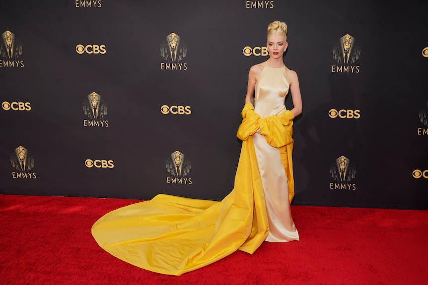 Anya wears a cream coloured floor-length silk dress, with a gigantic golden yellow wrap around her elbows