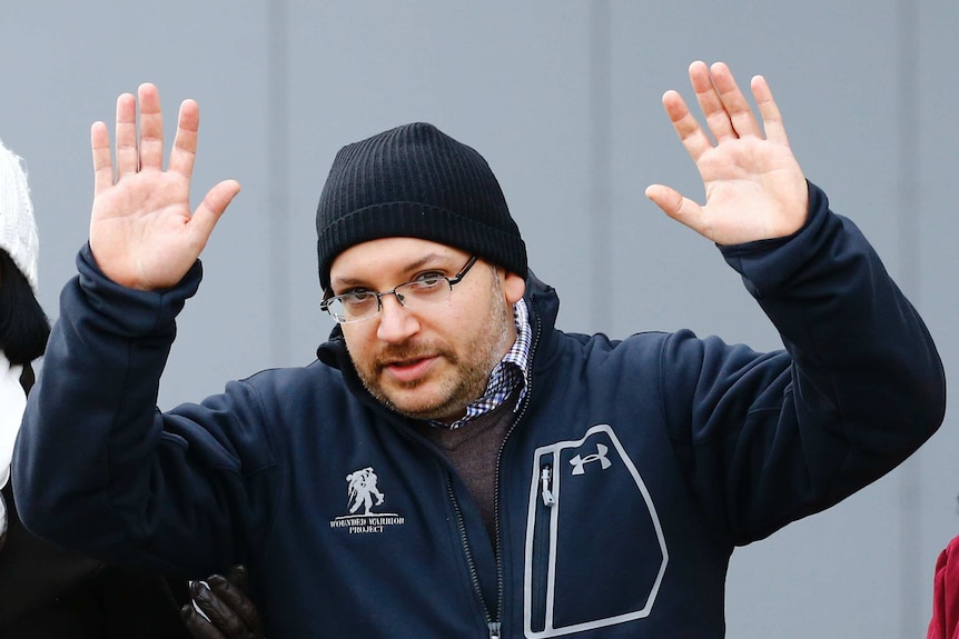 Jason Rezaian holds his hands in the air wearing warm clothing and a beanie.