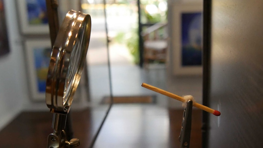 Close up photo showing a matchstick held in a clamp in front of a magnifying glass in an art gallery
