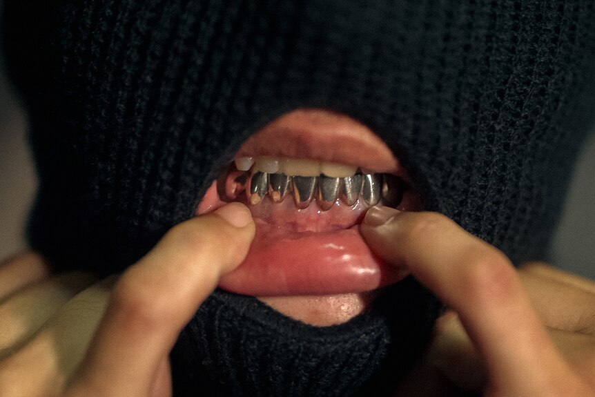 A woman pulls down her bottom lip to show silver grills covering her lower teeth. She wears a black balaclava.